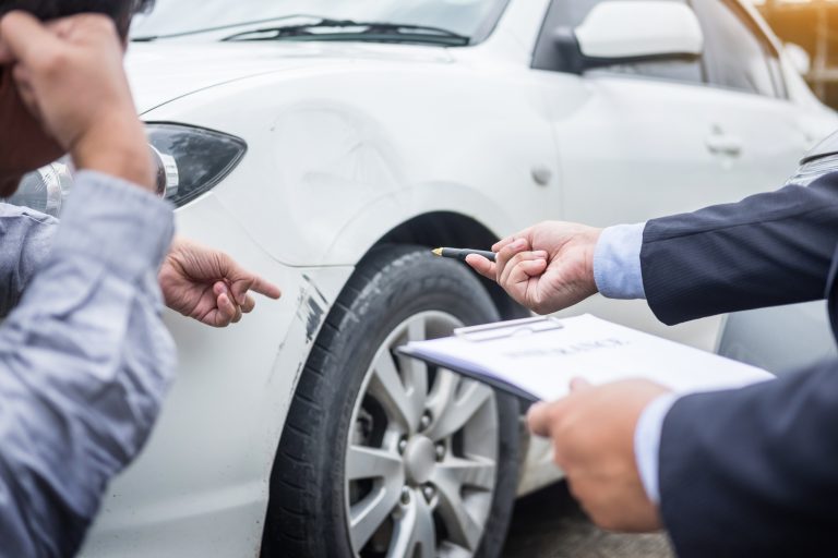 Insurance agent writing on clipboard while examining car after accident claim being assessed and processed.