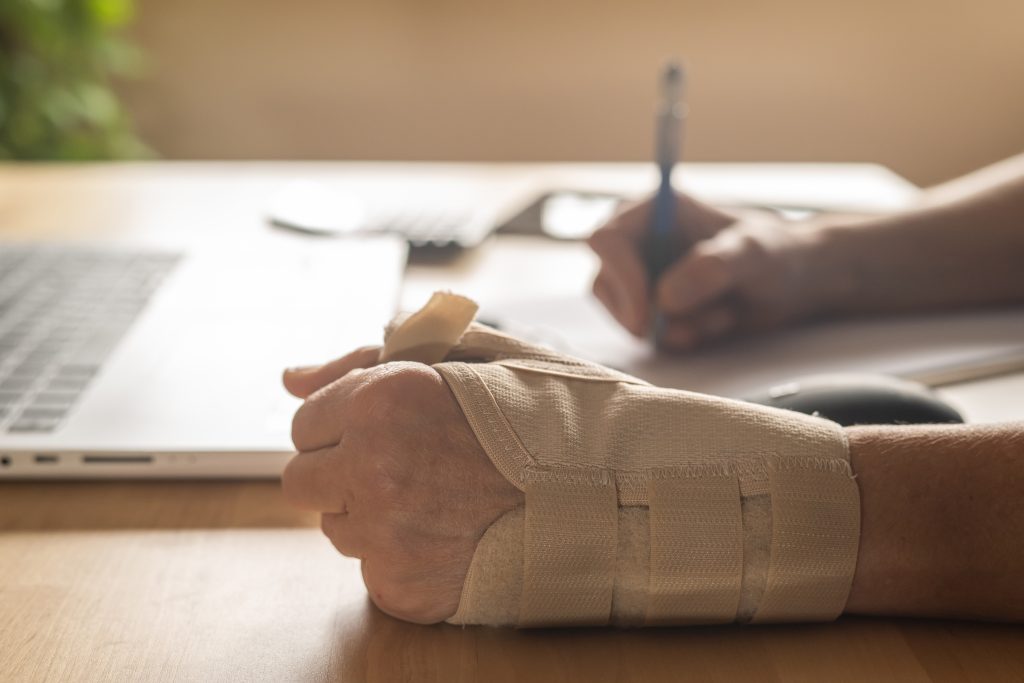 Professional Filling Out Workplace Injury Form Due To Wrist Pain From Repetitive Office Work, Illustrating Risks Of Carpal Tunnel And Tendonitis From Strain. Sick Leave