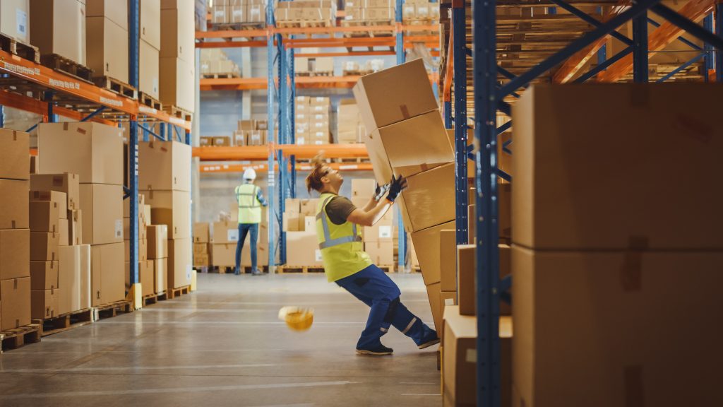 Shot of a Warehouse Worker Has Work Related Accident. He is Falling Down BeforeTrying to Pick Up Heavy Cardboard Box from the Shelf. Hard Injury at Work.
